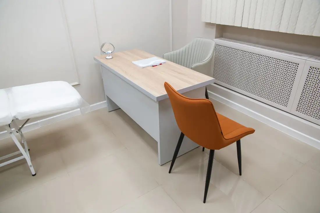A calm, bright consultation room with a desk, an orange chair and an exam bed
