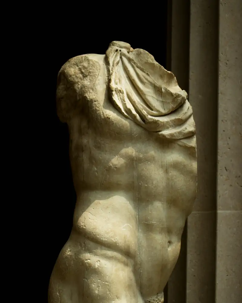 A classical marble torso in a dark gallery, lit from one side so the abdomen is in relief
